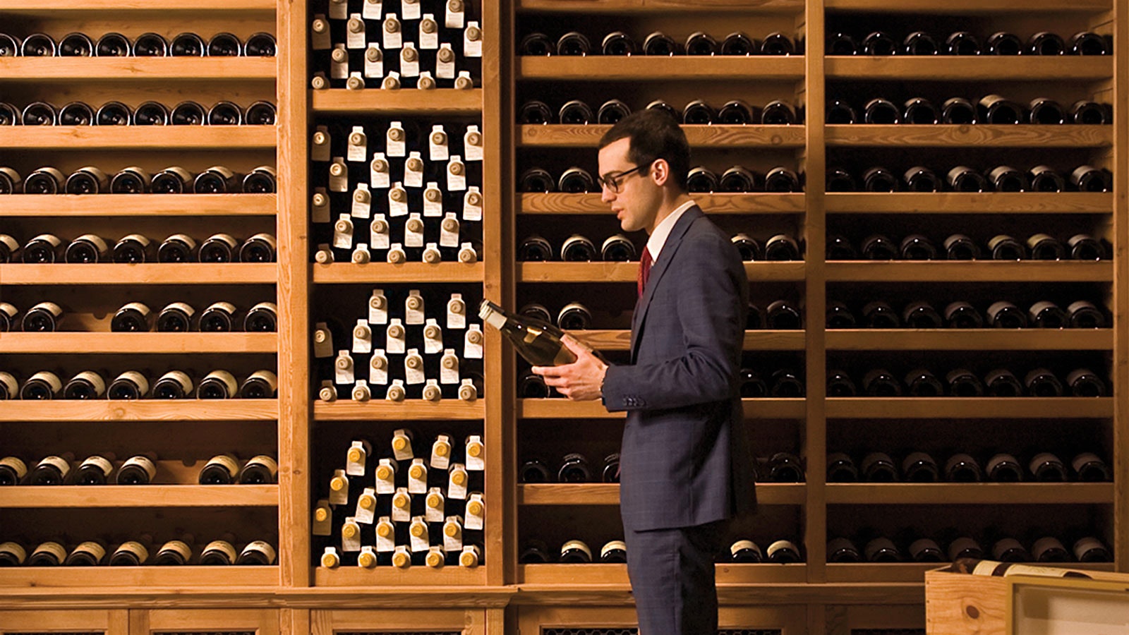   Cracco vyno direktorius, laikantis butelį priešais medinių vyno lentynų sieną, pilną butelių